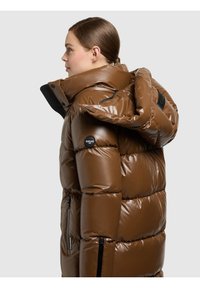 Ruda pūstinė striukė su blizgiu paviršiumi, dideliu gaubtu ir dvigubomis užtrauktukais. Yra juoda logotipo dėmė ir kontrastingos siūlės, suteikiančios struktūrą.