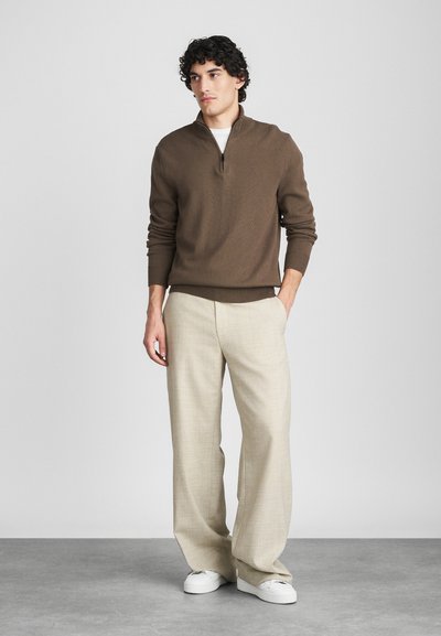 Pull en maille marron avec zip, pantalon large à carreaux beiges, baskets blanches, le modèle se tient devant un fond neutre.