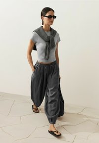 Femme portant des lunettes de soleil, un t-shirt gris moulant, un pantalon ample gris foncé, des sandales et un pull rayé posé sur les épaules, marchant à l'intérieur.