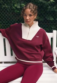 Bordozsarkana džemperis ar baltu rāvējslēdzēja imitācijas apkakli un V veida dizainu, kombinēts ar atbilstošām legingiem ar baltu jostasvietu.