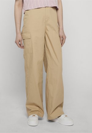 Osoba nosząca luźne beżowe spodnie cargo z klapami na kieszeniach oraz białe trampki, stojąca na jednolitym jasnym tle.