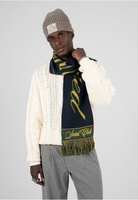 Sweter w kolorze kremowym z dzianymi wzorami, wełniana czapka, oraz granatowy szalik z żółtymi akcentami i frędzlami, na którym widnieje napis "Social Club."