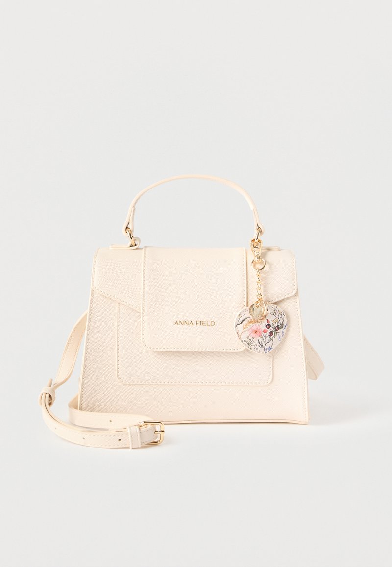 Μικρή κρεμ τσάντα χειρός με πάνω λαβή, ρυθμιζόμενο ιμάντα ώμου, εμπρόσθιο καπάκι με το λογότυπο «ANNA FIELD» και φλοράλ μπρελόκ σε σχήμα καρδιάς.