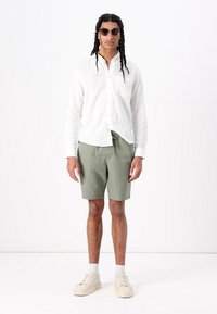 Chemise blanche en lin à manches longues, shorts verts, baskets légères, portés avec des lunettes de soleil. La chemise a une poche ; les shorts ont une taille élastique.