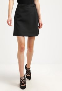 Jupe noire trapèze en tissu lisse avec un détail de ceinture fine à la taille. Associée à des chaussures à talons hauts noires à fines brides.