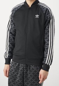 Černá sportovní bunda na zip se šedými rukávy s vzorem, vybavená třemi bílými pruhy a logem Adidas na hrudi.