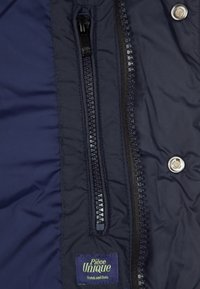 Nahaufnahme einer marineblauen Jacke mit zwei sichtbaren Reißverschlüssen, einem Druckknopf und einem kleinen Etikett mit der Aufschrift "Pièce Unique Scotch and Soda."