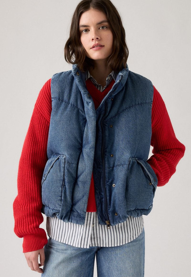 Denim puffer vest i blå farge, med kviltet tekstur og trykknapper. Brukt over en rød genser og stripete skjorte, med to sidelommer.