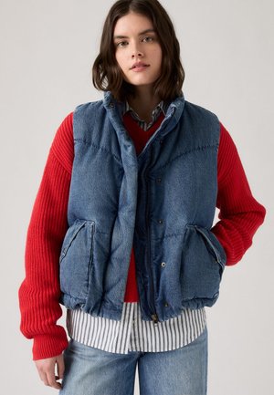 Gilet imbottito in denim di colore blu, con texture trapuntata e bottoni a scatto. Indossato sopra un maglione rosso e una camicia a righe, con due tasche laterali.