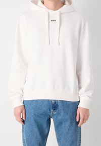 Personne portant un sweat à capuche blanc avec le texte « HUGO » sur la poitrine et un jean bleu, debout devant un fond clair uni.