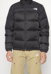 Czarna kurtka puchowa z pikowanym wzorem, wysokim kołnierzem, zamkiem z przodu i logiem na lewej piersi. Nosi na sobie beżowe szorty z kieszeniami.