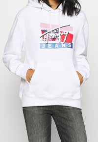 Biała hoodie wykonana z mieszanki bawełny z sznurkami. Posiada kolorowy nadruk z napisem "Tommy Jeans" oraz kieszeń kangurkę.