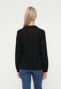 Black, long-sleeve top featuring intricate cut-out patterns and textured fabric. Back has a central slit and is designed for a loose fit.