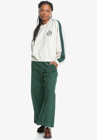 Sudadera de felpa crema con mangas verdes y logo, combinada con pantalones de pana anchos verdes y zapatos marrones con cordones.
