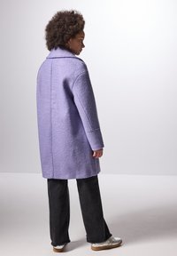 Cappotto di lana lilla con una texture, colletto sartoriale, maniche lunghe e spacco laterale, abbinato a pantaloni larghi scuri e scarpe argentate.