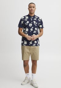 Kortermet marineblå polo med hvit blomstermønster, sammen med beige shorts. Sneakers og hvite sokker er brukt.