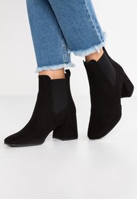 Botines de ante negro con tacones cuadrados, punteras afiladas y paneles elásticos laterales. Llevados con unos jeans cortos deshilachados de color azul claro.