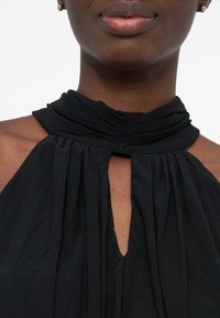 Κοντινό πλάνο ενός ατόμου που φοράει μαύρο τοπ με λαιμόκοψη halter, ραμμένο με μαζεμένο ύφασμα και μικρό κεντρικό άνοιγμα στο στήθος.