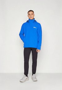 Mėlyna neperšlampama striukė su aukštu apykakliu, užtrauktuku ir reguliuojama gaubtu; derinama su tamsiomis kelnėmis ir šviesiai pilkais batais.