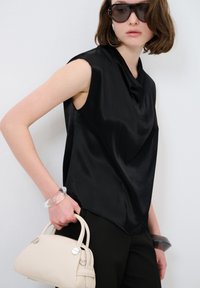 Top nero senza maniche in tessuto satinato con scollatura drappeggiata, abbinato a pantaloni neri. Gli accessori includono un bracciale trasparente e una borsa color crema.
