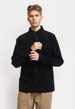 Zwarte corduroy overhemd met een knoopsluiting aan de voorkant, puntige kraag en twee borstzakken. Lange mouwen met knoopsluiting en een gestructureerde afwerking.