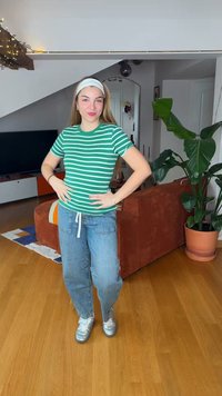 Young woman in green striped shirt and jeans stands in a living room with wooden floor, orange couch, large potted plant, and TV in background.
