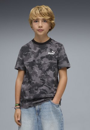 Ragazzo biondo che indossa una t-shirt Puma grigia con motivo mimetico e pantaloncini di jeans azzurri, con una mano in tasca, su sfondo grigio.