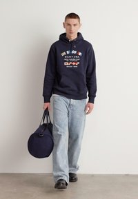 Felpa navy con grafiche, jeans azzurri larghi, e scarpe nere; porta una borsa da viaggio rotonda navy, su uno sfondo neutro.