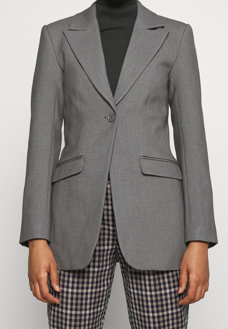 Blazer gris ajusté avec fermeture à un bouton, poches à rabat, porté sur un col roulé noir et un pantalon à carreaux.