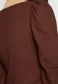 Brun texturerad blus med ett mjukt ribbat material, som har hopdragna detaljer vid axeln och en vid halsringning.