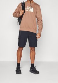 Sudadera marrón con logo blanco, pantalones cortos azul marino con bolsillos con cremallera y zapatillas negras. El conjunto combina comodidad y funcionalidad.