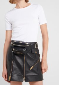 Žena v bílém tričku s krátkým rukávem, černé kožené sukni se zlatými zipy a černé kožené ledvince s zlatými detaily a logem.
