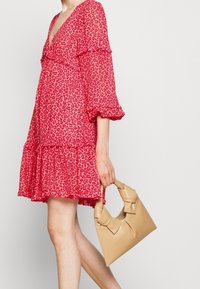 Donna che indossa un abito rosso con stampa leopardata e maniche a volant, mentre tiene una borsa di pelle beige con manici annodati.