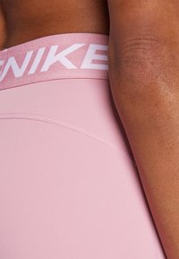 Világos rózsaszín sportleggings vastag derékrésszel, amelyen a "NIKE" felirat található, sima anyagból készült, és laposvarrásos részletekkel rendelkezik.