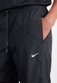 Črne Nike kratke hlače iz lahkega materiala, z elastičnim pasom, vrvico, stranskimi žepi in belim logotipom na levem stegnu.