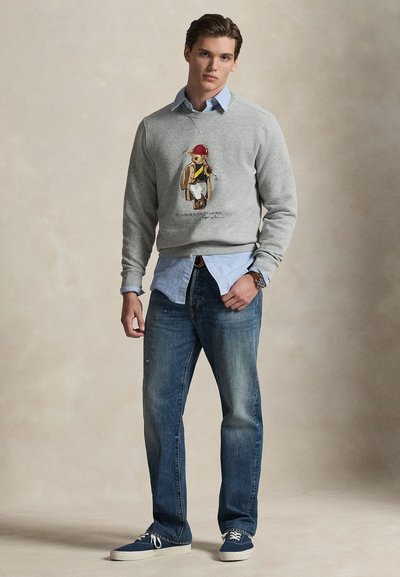 Γκρι φούτερ με γραφικό τύπωμα αρκούδας με ρούχα, σε συνδυασμό πάνω από ένα ανοιχτό μπλε πουκάμισο με γιακά, μαζί με μπλε τζιν και αθλητικά παπούτσια.