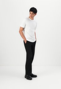 T-shirt blanc à manches courtes, pantalon noir et chaussures noires. Design simple, texture lisse, avec une coupe décontractée. Mains dans les poches, regard tourné vers le bas.