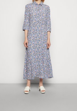 Femme portant une robe midi à fleurs bleues avec col et manches trois-quarts, associée à des sandales blanches à talons blocs et bout ouvert.