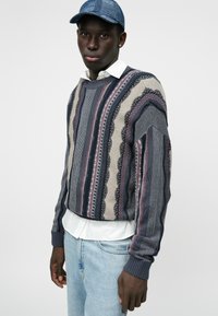 Πλεκτό πουλόβερ σε ναυτικό, κρέμ και μπορντώ με κατακόρυφες ρίγες και υφασμάτινα σχέδια; στρογγυλή λαιμόκοψη και ριμπ μανσέτες. Συνδυασμένο με ένα λευκό πουκάμισο.