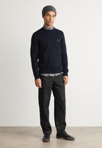 Dark blue sweater with crew neck and small logo, layered over a light blue button-up shirt. Paired with dark denim jeans and black sneakers.