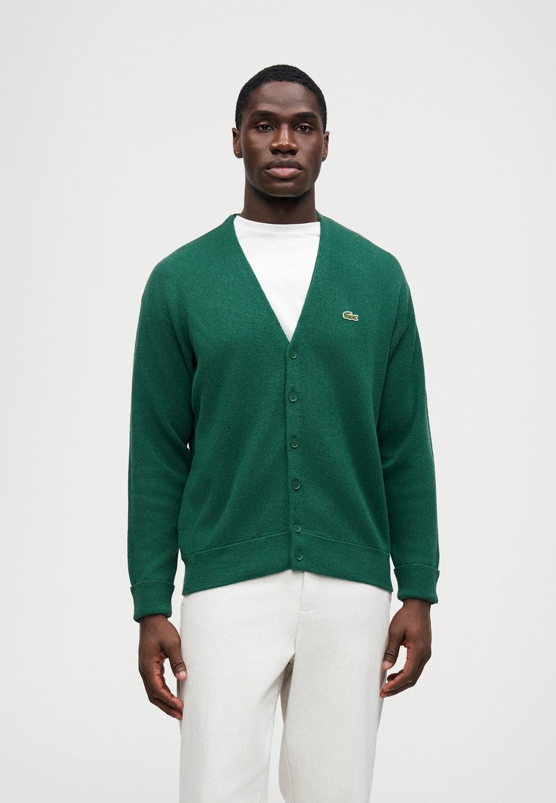 Cárdigan verde de punto con escote en V, que cuenta con seis botones y un logo sutil en el lado izquierdo del pecho. Se lleva sobre una camiseta blanca.