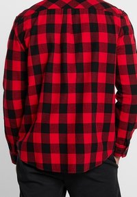 Camicia in flanella a quadri rossi e neri con maniche lunghe, polsini con bottoni e una piega sul retro per un comfort aggiuntivo. Texture liscia, design casual.