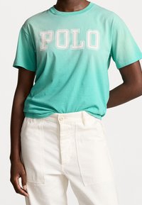 Světle tyrkysové bavlněné tričko s velkým bílým nápisem "POLO". Krátké rukávy a volný střih v kombinaci s bílými kalhotami. Hladká textura.