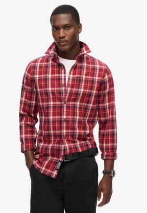 Camisa de cuadros rojos con acentos blancos y azul marino, mangas largas, cuello abotonado y una etiqueta en el dobladillo, combinada con pantalones negros y un reloj.