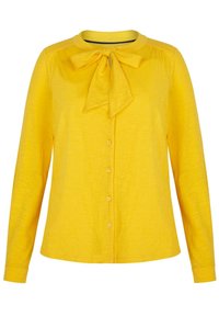 Blusa amarilla de manga larga hecha de una tela suave, con un lazo en el cuello y cierre frontal de botones a juego.