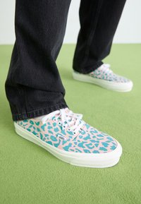 Canvas sneakers with a pink base and blue leopard print design, white laces, and a white rubber sole, worn with black denim pants.