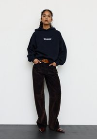 Granatowa bluza z kapturem z białym napisem, luźny krój, w zestawie z ciemnymi szerokimi spodniami sztruksowymi i butami z zaostrzonym noskiem. Brązowy pasek podkreśla talię.