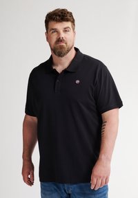 Mustapolo, jossa on kaulus ja lyhyet hihat, vasemmassa rinnassa on pieni pyöreä logo. Materiaali vaikuttaa sileältä ja kevyeltä.