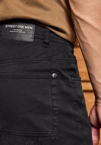 Zbliżenie osoby w czarnych dżinsach z metką "Street One Men" oraz brązowej koszuli, koncentrując się na tylnej kieszeni i szwie.