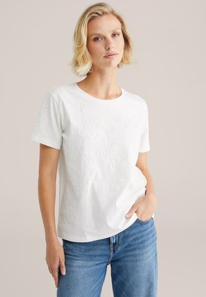 Valkoinen puuvillapaita, jossa on teksturoitua kukka-embroideryä edessä, pyöreä kaula-aukko, lyhyet hihat ja rento istuvuus.